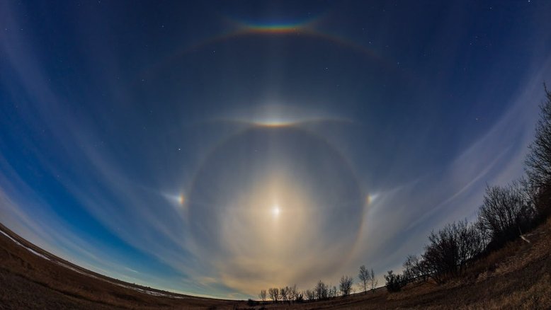 Uma Lua quase na fase Cheia, em abril de 2022, fez esta aparição luminosa para uma noite de primavera, no Norte de Alberta, no Canadá. O luar brilhante refratado e refletido por cristais de gelo hexagonais em nuvens altas criou um complexo de halos e arcos mais comumente vistos pela luz solar nos céus diurnos. Com Arcturus, a estrela Alfa da Constelação de Bootes (“O Boieiro”), à esquerda, e Regulus, a principal estrela da Constelação de Leão, à direita, a Lua está centrada em seu halo de 22 graus