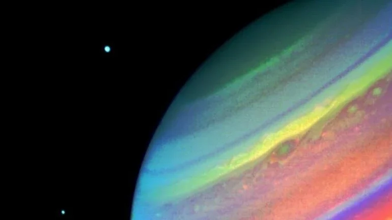 Nesta imagem de Saturno, feita pela espaçonave Voyager 2, em agosto de 1981, é possível ver os seus satélites Dione, à direita, e Encélado, à esquerda. Este registro em cores falsas mostra características da atmosfera do planeta. Muitas nuvens convectivas aparecem entre as duas manchas, que são típicas da região. O projeto Voyager 2 lançou ao espaço a sonda de mesmo nome, em 1977, sendo gerenciado pela NASA e pelo Jet Propulsion Laboratory (JPL), a partir de sua sede, em Pasadena, na Califórnia. O equipamento ainda permanece em contato com a NASA, já transitando pelos limites do Sistema Solar