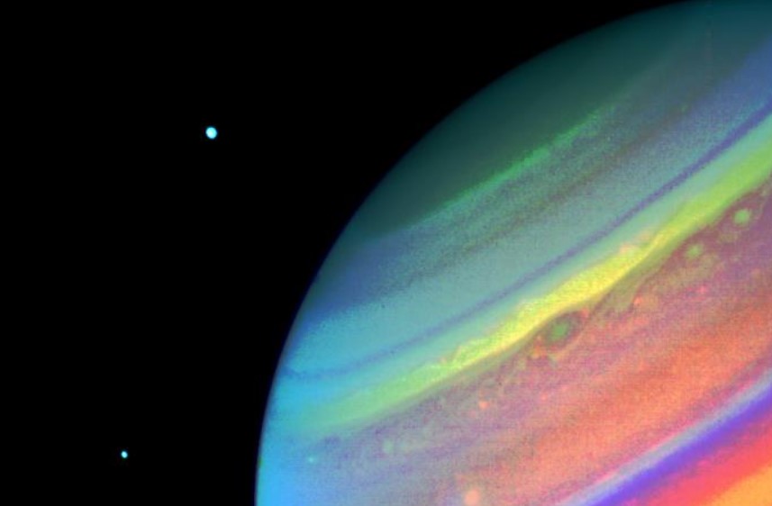 Nesta imagem de Saturno, feita pela espaçonave Voyager 2, em agosto de 1981, é possível ver os seus satélites Dione, à direita, e Encélado, à esquerda. Este registro em cores falsas mostra características da atmosfera do planeta. Muitas nuvens convectivas aparecem entre as duas manchas, que são típicas da região. O projeto Voyager 2 lançou ao espaço a sonda de mesmo nome, em 1977, sendo gerenciado pela NASA e pelo Jet Propulsion Laboratory (JPL), a partir de sua sede, em Pasadena, na Califórnia. O equipamento ainda permanece em contato com a NASA, já transitando pelos limites do Sistema Solar