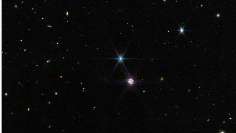 Nesta imagem captada pelo Telescópio Espacial James Webb, vê-se algumas centenas de galáxias de fundo ao lado do sistema formado pelo planeta Netuno e seus 14 satélites naturais. Além do gigante gasoso, retratado com os seus anéis, domina a imagem um ponto de luz muito brilhante, com picos de difração, que não é uma estrela, mas a maior de suas luas, Tritão. Netuno, quando comparado à Terra, é um planeta gigante. Se a Terra fosse do tamanho de uma moeda de cinco centavos, Netuno seria tão grande quanto uma bola de basquete. No entanto, Netuno parece relativamente pequeno nesta visão de campo ampla de nosso vasto universo