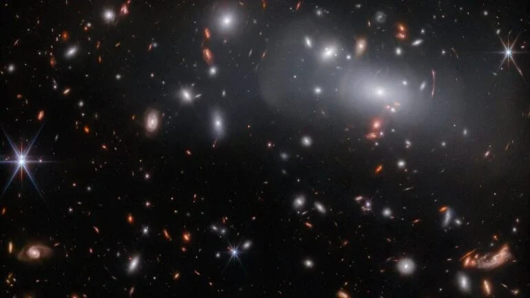 Esta imagem do Telescópio Espacial James Webb apresenta o enorme aglomerado de galáxias RX J2129. Devido às lentes gravitacionais, esta observação contém três imagens diferentes da mesma galáxia que hospeda uma supernova. As lentes gravitacionais ocorrem quando um corpo celeste massivo causa uma curvatura suficiente do espaço-tempo para dobrar o caminho da luz. Neste caso, a lente é o aglomerado de galáxias RX J2129, localizado a cerca de 3,2 bilhões de anos-luz da Terra, na Constelação de Aquário. As lentes gravitacionais podem fazer com que os objetos de fundo fiquem distorcidos, como pode ser visto pelos arcos concêntricos de luz no canto superior direito