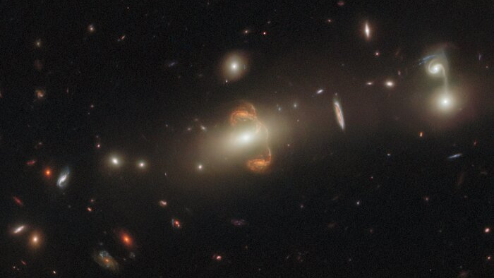 Esta imagem do Telescópio Espacial Hubble mostra a galáxia SGAS J143845+145407 sob o efeito de lentes gravitacionais. O fenômeno resultou numa imagem espelhada da galáxia ao centro, criando uma peça cativante. As lentes gravitacionais ocorrem quando um corpo celeste massivo – como um aglomerado de galáxias – causa uma curvatura suficiente do espaço-tempo para que o caminho da luz ao seu redor seja visivelmente dobrado. As lentes gravitacionais podem resultar em múltiplas imagens, no objeto de fundo aparecendo como um arco distorcido ou mesmo um anel. Outra consequência importante desta distorção é a ampliação, permitindo a observação de objetos muito distantes ou tênues para serem vistos