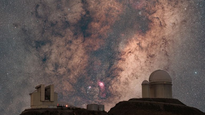 O núcleo da Via Láctea eleva-se ao fundo desta paisagem que inclui o Observatório de La Silla, no Chile. Ao centro da imagem de céu noturno profundo, vê-se o centro da nossa galáxia natal, a Via Láctea, na direção da Constelação de Sagitário. À esquerda, está o New Technology Telescope do Observatório Europeu do Sul (ESO) e, à direita, está o telescópio do ESO que é sede dos espectrógrafos caçadores de exoplanetas HARPS e NIRPS. Entre eles, o bojo central da galáxia está repleto de nuvens escuras de poeira interestelar, estrelas brilhantes, aglomerados e nebulosas. A proeminente emissão de hidrogênio avermelhado revela a Nebulosa Laguna (M8), com sua rica formação de estrelas. A Nebulosa Trífida (M20), que combina a luz azul de uma nebulosa de reflexão com a emissão avermelhada, está à esquerda de M8