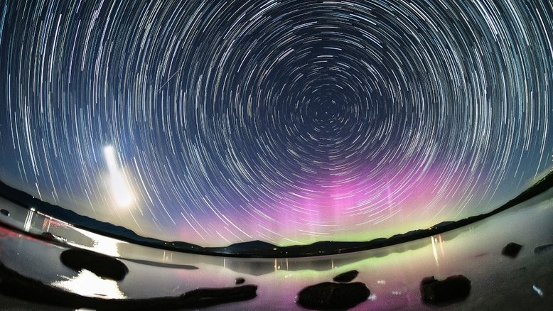 Arcos de trilhas estelares refletem a rotação diária do planeta Terra, nesta colorida paisagem noturna. Para criar a composição, em maio deste ano, foram registradas exposições consecutivas com uma câmera fixada em um tripé, nas margens do reservatório Ashokan, na região de Catskills, em Nova York, EUA. A estrela polar, Polaris, está perto do centro dos arcos da trilha estelar. O amplo rastro da Lua Crescente está à esquerda, lançando um forte reflexo nas águas do reservatório. Com a intensa atividade solar impulsionando as recentes tempestades geomagnéticas, a colorida Aurora Boreal, rara na região, brilha sob a Pólo Celeste Norte
