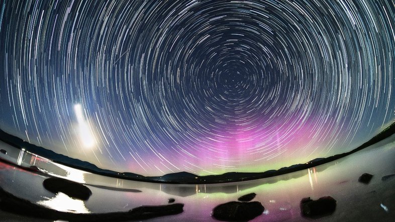 Arcos de trilhas estelares refletem a rotação diária do planeta Terra, nesta colorida paisagem noturna. Para criar a composição, em maio deste ano, foram registradas exposições consecutivas com uma câmera fixada em um tripé, nas margens do reservatório Ashokan, na região de Catskills, em Nova York, EUA. A estrela polar, Polaris, está perto do centro dos arcos da trilha estelar. O amplo rastro da Lua Crescente está à esquerda, lançando um forte reflexo nas águas do reservatório. Com a intensa atividade solar impulsionando as recentes tempestades geomagnéticas, a colorida Aurora Boreal, rara na região, brilha sob a Pólo Celeste Norte