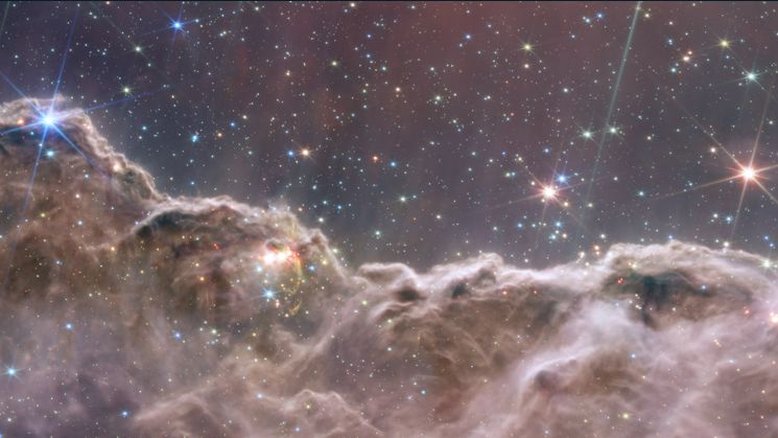 As câmeras do Telescópio Espacial James Webb produziram esta imagem inédita de uma das regiões de formação estelar na Nebulosa Carina, que fica na direção da Constelação de Carina. O que se parece com montanhas em uma noite de luar é, na verdade, a borda de uma jovem região de formação de estrelas, conhecida como NGC 3324. Essa borda de uma gigantesca cavidade gasosa fica a cerca de 7,6 mil anos-luz de distância. A área cavernosa foi esculpida pela intensa radiação ultravioleta e os ventos estelares de estrelas jovens extremamente massivas e quentes