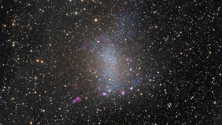 NGC 6822, também conhecida como Galáxia de Barnard, está na vizinhança da Via Láctea. Em meio à Constelação de Sagitário, ela fica a apenas 1,5 milhões de anos-luz de distância, sendo um membro do nosso Grupo Local de galáxias. Como galáxia anã irregular, NGC 6822 tem cerca de 7 mil anos-luz de diâmetro. As estrelas mais brilhantes e com uma aparência pontiaguda, que parecem em primeiro plano na imagem, são da nossa Via Láctea. Atrás delas, a Galáxia de Barnard aparece repleta de jovens estrelas azuis, exibindo também o brilho rosado do hidrogênio proveniente das regiões de formação estelar