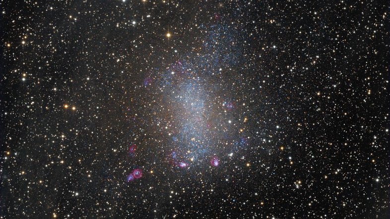 NGC 6822, também conhecida como Galáxia de Barnard, está na vizinhança da Via Láctea. Em meio à Constelação de Sagitário, ela fica a apenas 1,5 milhões de anos-luz de distância, sendo um membro do nosso Grupo Local de galáxias. Como galáxia anã irregular, NGC 6822 tem cerca de 7 mil anos-luz de diâmetro. As estrelas mais brilhantes e com uma aparência pontiaguda, que parecem em primeiro plano na imagem, são da nossa Via Láctea. Atrás delas, a Galáxia de Barnard aparece repleta de jovens estrelas azuis, exibindo também o brilho rosado do hidrogênio proveniente das regiões de formação estelar