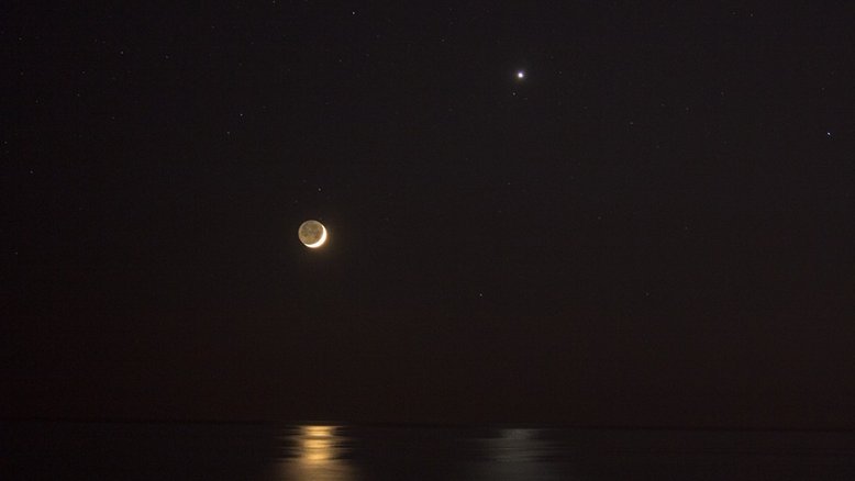 Posando perto do horizonte Oeste, o planeta Vênus aparece em sua versão de brilhante Estrela Vespertina, ao lado da Lua Nova jovem e esguia.  A dupla compartilhou reflexos em um mar calmo, após o pôr do Sol. Registrada em uma praia em Santa Marinella, perto de Roma, Itália, esta imagem exibe a adorável conjunção celestial dos dois objetos mais brilhantes no céu noturno. Também presente na foto, vê-se que a luz refletida da Terra ilumina o lado noturno lunar, em um fenômeno conhecido como Luz Cinérea 