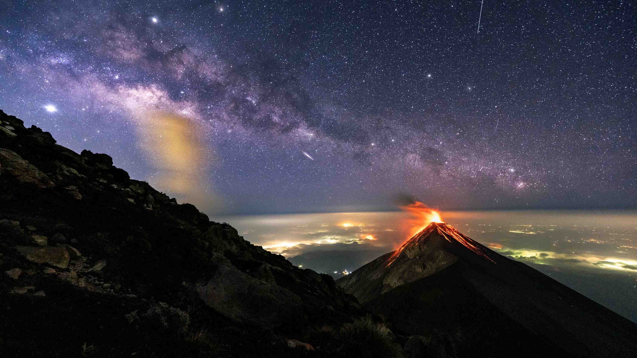 Nesta imagem, o Volcán de Fuego está em erupção, adornado por cinzas incandescentes e fluxos de lava. Na paisagem, localizada na Guatemala, acima das luzes das cidades vizinhas, vê-se o plano central da Via Láctea, a nossa galáxia. O planeta Júpiter também aparece no canto superior esquerdo, com a estrela brilhante Antares, logo à sua direita. O registro, feito em abril de 2019, foi capturado a partir da lateral do vulcão Acatenango, que fica na mesma região