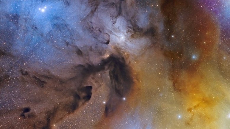 Por que o céu perto de Antares, a estrela Alfa da Constelação de Escorpião, e Rho Ophiuchi é tão empoeirado e colorido? As cores resultam de uma mistura de objetos e processos. A poeira fina – iluminada pela luz das estrelas – produz nebulosas de reflexão azuis. Nuvens gasosas, cujos átomos são excitados pela luz ultravioleta das estrelas, produzem nebulosas de emissão avermelhadas. Nuvens de poeira iluminadas bloqueiam a luz das estrelas e parecem escuras. Antares, a supergigante vermelha que é uma das estrelas mais brilhantes do céu noturno, ilumina as nuvens amarelo-avermelhadas, no canto inferior direito desta imagem. O sistema estelar Rho Ophiuchi fica no centro da nebulosa de reflexão azul, no canto superior esquerdo