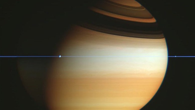 Quando os anéis de Saturno desapareceram, em 1612, Galileu não entendeu por quê. Mais tarde, naquele século, entendeu-se que as saliências incomuns de Saturno eram anéis e que, quando a Terra cruza o seu plano, eles parecem desaparecer. Isso ocorre porque os anéis de Saturno estão confinados a um plano muito mais fino. Esta imagem foi feita pela missão Cassini, enquanto a espaçonave robótica cruzava o plano dos anéis de Saturno. O fino plano está em azul, faixas e nuvens na atmosfera superior de Saturno aparecem em dourado e as sombras escuras dos anéis curvam-se no topo do planeta gigante gasoso. As luas do planeta são mostradas como saliências nos anéis
