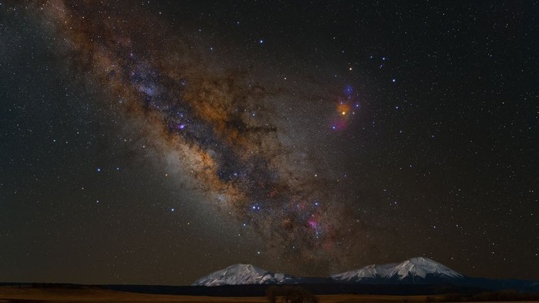 A faixa diagonal que domina esta imagem é a visão de um dos braços da Via Láctea, com destaque à região central da nossa galáxia. A paisagem retrata os Picos Espanhóis, localizados no Colorado, EUA. À direita da empoeirada Via Láctea, a grande região colorida rodeia a estrela Rho Ophiuchi, da Constelação do Serpentário. Os planetas Marte e Saturno estão visíveis acima dos picos de rocha, formando um triângulo laranja junto com a brilhante Antares, a estrela Alfa da Constelação de Escorpião