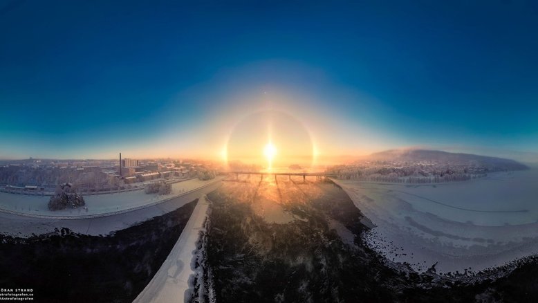 Eis uma visão do Sol próximo ao Solstício de dezembro, que marca o início astronômico do verão, no Hemisfério Sul, e do inverno, no Hemisfério Norte. Nesta imagem, registrada em dezembro de 2022, o Sol estava logo acima do horizonte ao meio-dia local de Ostersund, no centro da Suécia. A vista sobre a cidade à beira do lago exibe o Sol do meio-dia com um belo halo solar de gelo. Cristais de gelo atmosféricos que ocorrem naturalmente produzem o halo, ao refratar e refletir a luz solar através de sua geometria hexagonal. Ainda assim, com o Sol baixo e perto do horizonte no céu claro, entre as ​​fontes dos cristais de gelo que produzem este halo intenso estão os canhões de neve. Operando em uma área de esqui local, as máquinas de fazer neve criam uma pluma visível no topo da ilha vizinha, Froson, no lado direito do panorama