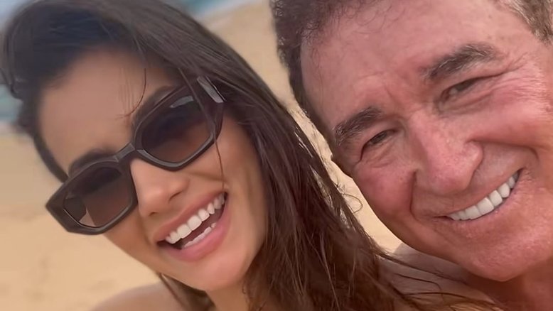 O cantor Amado Batista e a modelo Calita Franciele Miranda de Souza aparecem em uma selfie, com os rostos fotografados bem de perto. Ao fundo, vê-se o mar