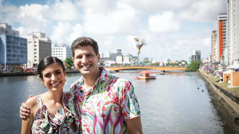 Tabata Amaral e João Campos aparecem com roupas coloridas. Ao fundo, vemos o bloco de carnaval Galo da Madrugada