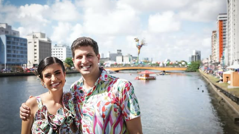 Tabata Amaral e João Campos aparecem com roupas coloridas. Ao fundo, vemos o bloco de carnaval Galo da Madrugada