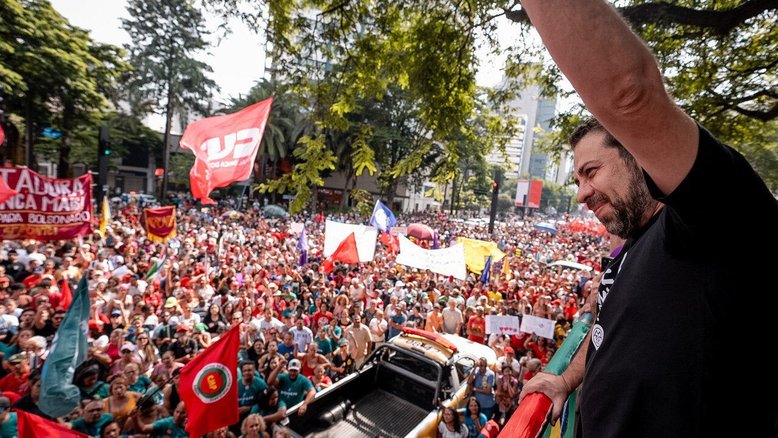 Guilherme Boulos está no topo de um carro elétrico com um punho para o alto. Ao redor, é possível ver pessoas com faixas e bandeiras do protesto "Sem Anistia"