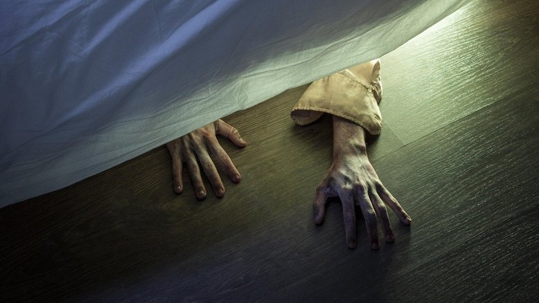 A fotografia mostra de cima a lateral de uma cama. Podemos pelo ângulo ver duas mãos de alguém embaixo da cama