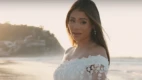 A cantora Lexa aparece com um vestido branco de renda em uma praia. Sua boca está aberta, como se estivesse cantando ao ser fotografada