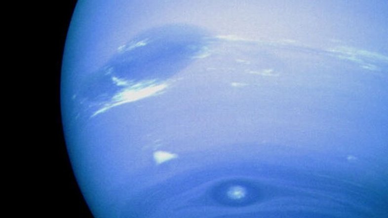Netuno tem manchas. O gigante gasoso mais externo do Sistema Solar apresenta uma tonalidade azul quase uniforme, criada por pequenas quantidades de metano à deriva em uma atmosfera espessa de hidrogênio e hélio quase incolores. Há também manchas escuras que são anticiclones: grandes sistemas de alta pressão que giram no topo das nuvens frias de Netuno. Nesta imagem, tirada pela sonda espacial robótica Voyager 2, em 1989, vê-se o que foi denominada sua Grande Mancha Escura, do tamanho da Terra (à extrema esquerda), e a Mancha Escura 2, perto da parte inferior