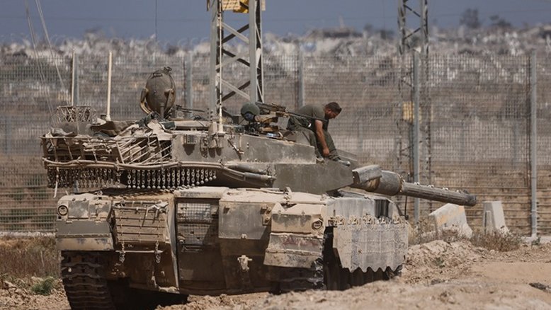 Tanque israelense na fronteira de Gaza. Ao fundo, fumaça e destroços aparecem desfocados. Imagem registrada em 27/05/2025, um ano e meio após o ataque do Hamas que iniciou um conflito com mais de 55 mil mortos, a maior parte de palestinos