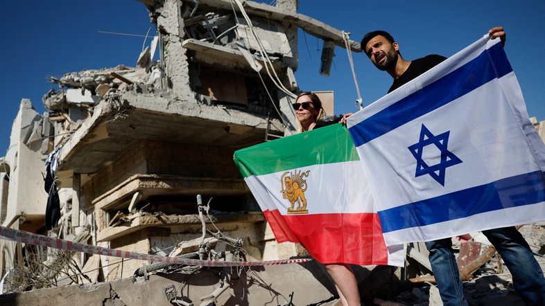 Pessoas seguram uma antiga bandeira real iraniana e uma bandeira israelense no local do impacto após ataque de mísseis do Irã contra Israel, em Rishon LeZion, Israel, em 14 de junho de 2025