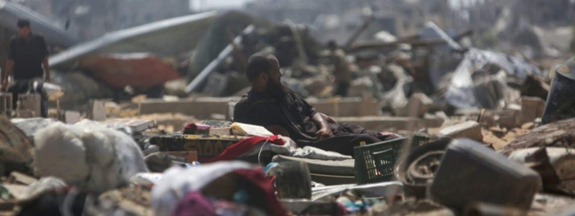 Um homem deitado sobre objetos recuperados observa palestinos inspecionarem barracas de um campo de deslocados que foram destruídas por bombardeios israelenses, perto de Khan Yunis, no sul da Faixa de Gaza, em 11 de julho de 2025 - AFP